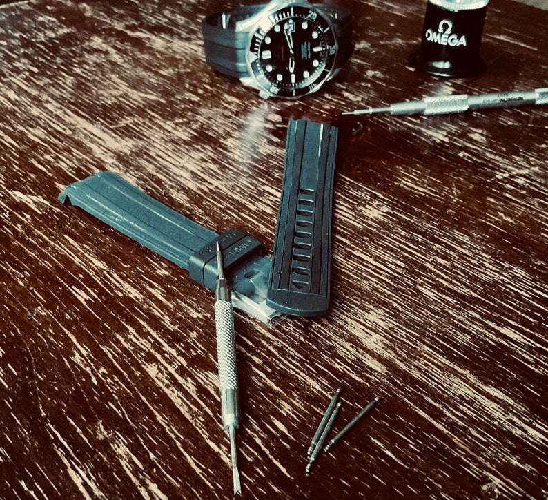 May include: A close-up of a black watch band with a silver watch face and silver watch hands. The watch band is laying on a wooden surface with a silver watch tool and a small bottle of oil labeled "OMEGA".
