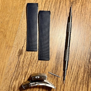 Puede incluir: Dos correas de reloj negras con una superficie texturizada y una correa de reloj plateada con una herramienta de pasador de resorte.