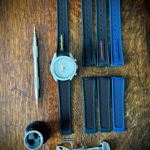 Peut inclure: Une montre-bracelet avec un cadran bleu clair et des bracelets noirs et bleus, ainsi que des outils et accessoires. Les bracelets de montre portent l'inscription "Speedmaster". Les articles sont disposés sur une surface en bois.