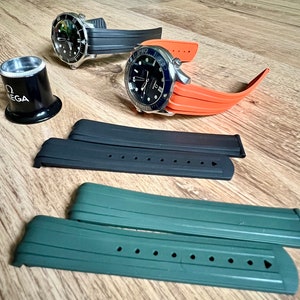 Puede incluir: Dos correas de reloj negras y naranjas con una lupa. Las correas de reloj son de goma y tienen una superficie texturizada. La lupa es de metal y tiene un mango negro.