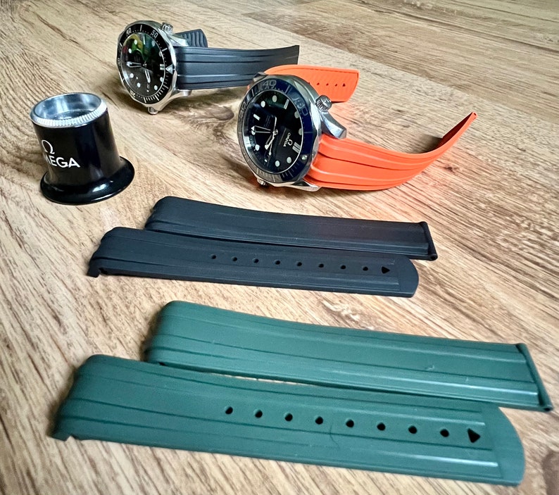 Puede incluir: Dos correas de reloj negras y naranjas con una lupa. Las correas de reloj son de goma y tienen una superficie texturizada. La lupa es de metal y tiene un mango negro.