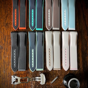 May include: A collection of watch straps in various colors, including black, navy, brown, and light blue, with textured surfaces. The straps are arranged on a wooden surface, along with watch repair tools and a clasp.