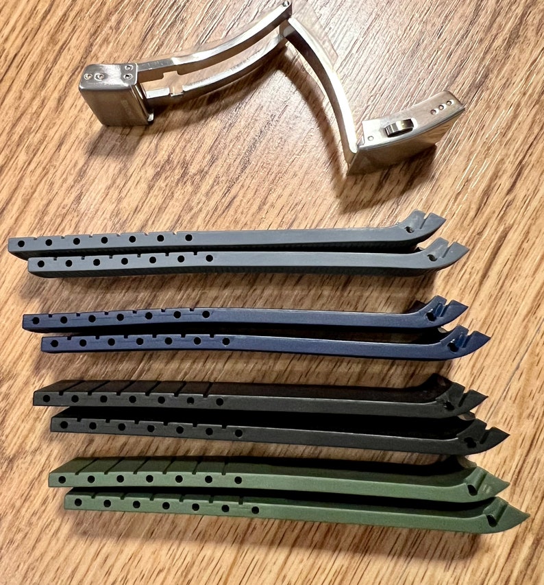 Puede incluir: Cuatro correas de reloj en diferentes colores: negro, azul oscuro, verde y gris. Las correas son de goma y tienen una superficie texturizada. Tambi&eacute;n se muestra una hebilla de reloj plateada.