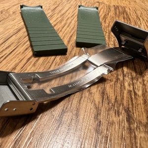 Puede incluir: Una hebilla de correa de reloj de plata con el texto "316L STAINLESS" y "316L STEEL" grabado en el metal. La hebilla est&aacute; unida a dos correas de reloj de goma verde.