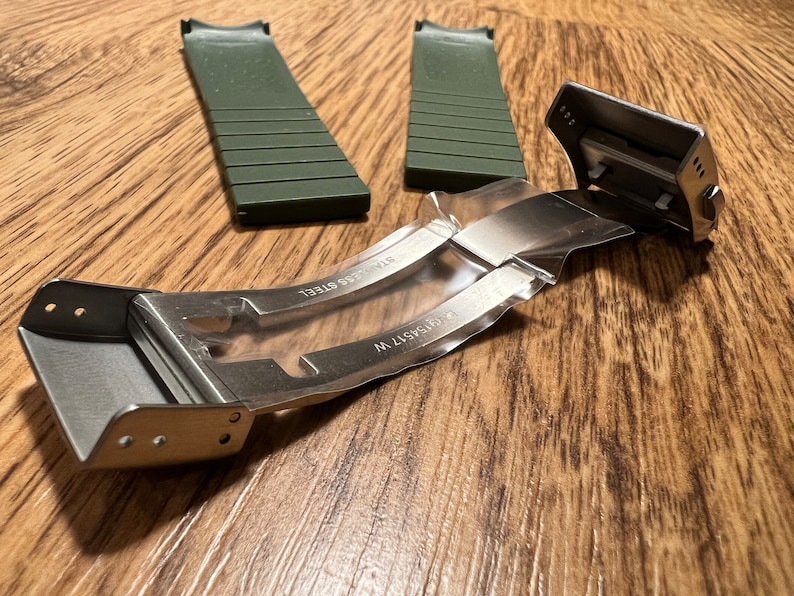 Puede incluir: Una hebilla de correa de reloj de plata con el texto "316L STAINLESS" y "316L STEEL" grabado en el metal. La hebilla est&aacute; unida a dos correas de reloj de goma verde.