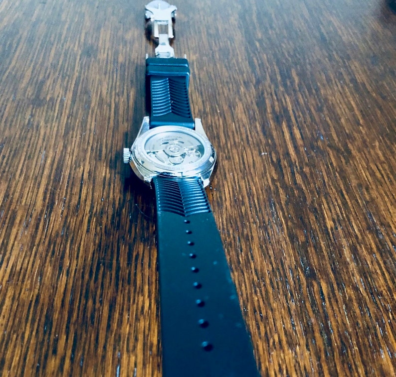 Puede incluir: Un reloj plateado con una correa de goma negra. La esfera del reloj es visible y muestra el funcionamiento interno del reloj.