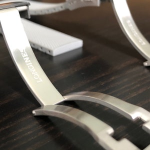 Puede incluir: Una correa de reloj plateada con la palabra "LONGINES" grabada en el cierre.