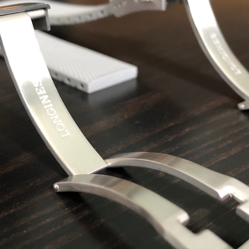 Puede incluir: Una correa de reloj plateada con la palabra "LONGINES" grabada en el cierre.