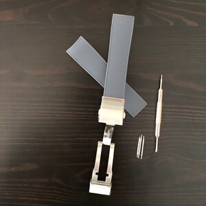 Puede incluir: Una correa de reloj de silicona gris con una hebilla plateada y pasadores de resorte. La correa tiene un patr&oacute;n texturizado.