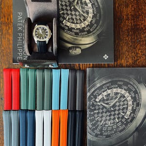 May include: A high-end wristwatch with a black strap is presented in a leather case, with a selection of vibrant watch straps. The watch features a silver dial and is shown with a book bearing the words "Patek Philippe".