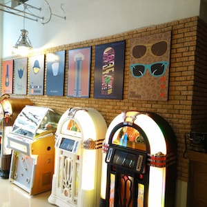 May include: Five vintage jukeboxes in a row, with a brick wall behind them. The jukeboxes are in various colors, including red, white, and black. The wall is decorated with posters, including one with the text "True Romance".