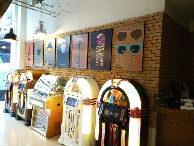 May include: Five vintage jukeboxes in a row, with a brick wall behind them. The jukeboxes are in various colors, including red, white, and black. The wall is decorated with posters, including one with the text "True Romance".