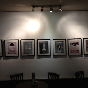 May include: A wall display of eight framed black and white posters with various designs. The posters are hung in a row on a white wall. The posters are all the same size and are framed in black.