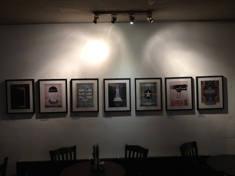 May include: A wall display of eight framed black and white posters with various designs. The posters are hung in a row on a white wall. The posters are all the same size and are framed in black.