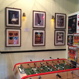 May include: A vintage foosball table with red and white accents, surrounded by framed posters and flyers. The posters feature various designs, including a pair of sunglasses, a hand, and a vintage camera. The flyers advertise events with text in Spanish and English.