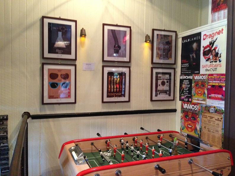 May include: A vintage foosball table with red and white accents, surrounded by framed posters and flyers. The posters feature various designs, including a pair of sunglasses, a hand, and a vintage camera. The flyers advertise events with text in Spanish and English.