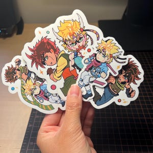 May include: A collection of four die-cut vinyl stickers featuring anime-style characters with spiky hair in various colours including brown, blonde, and blue. The illustrations depict characters in dynamic poses with colourful outfits and small star accents.