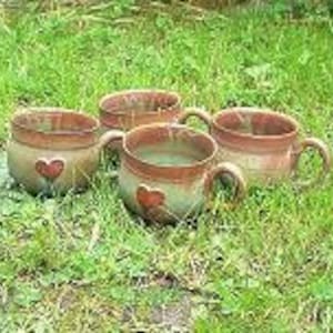 May include: Four brown ceramic mugs with heart-shaped cutouts and handles. The mugs are sitting on a bed of green grass.