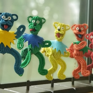 May include: Five colorful crocheted bear figurines, each with a different color, standing on a black wooden base. The bears are arranged in a line from left to right, with the blue bear in the front, followed by the green, yellow, orange, and red bears.