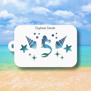 May include: A white rectangular stencil with rounded corners featuring a cutout design of a mermaid in the center, flanked by two seashells, two starfish, four small bubbles, and four small four-pointed stars. The text Daydream Stencils is printed.