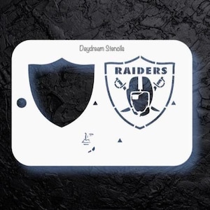 May include: White stencil featuring two shield designs. One shield is a solid shape, and the other displays the word "RAIDERS" above a helmet and crossed swords. The text "Daydream Stencils" is at the top.