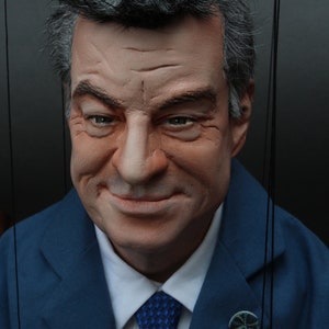Könnte beinhalten: Ein Mann in einem blauen Anzugjackett mit weißem Hemd und einer blauen gemusterten Krawatte. Er trägt eine kleine Anstecknadel am Revers seiner Jacke.