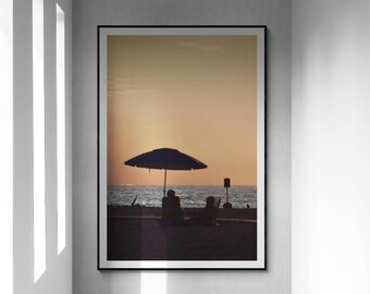May include: A framed photograph of a beach scene with two people sitting under a blue umbrella at sunset. The sky is a warm orange and yellow color.