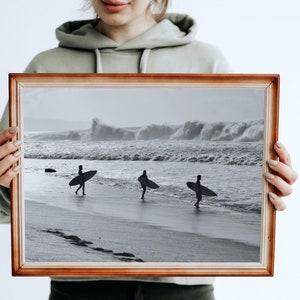May include: A framed black and white photograph of three surfers walking on a beach with their surfboards. The ocean is in the background with large waves crashing.