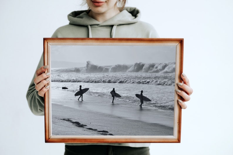 May include: A framed black and white photograph of three surfers walking on a beach with their surfboards. The ocean is in the background with large waves crashing.