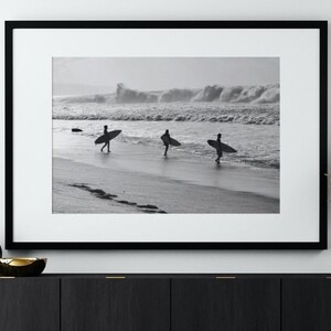 May include: Black and white photograph of three surfers walking on a beach with their surfboards. The surfers are silhouetted against the waves in the background.