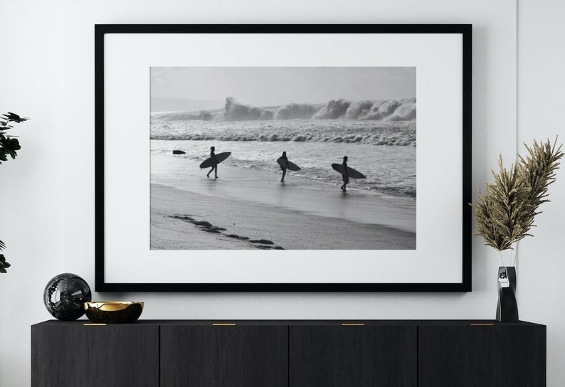 May include: Black and white photograph of three surfers walking on a beach with their surfboards. The surfers are silhouetted against the waves in the background.