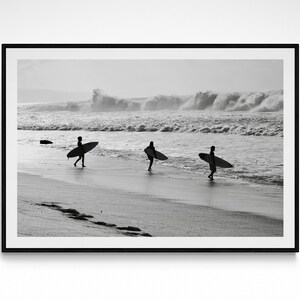 May include: Black and white photograph of three people walking on a beach with surfboards in hand. The people are silhouetted against the ocean and the waves crashing in the background.