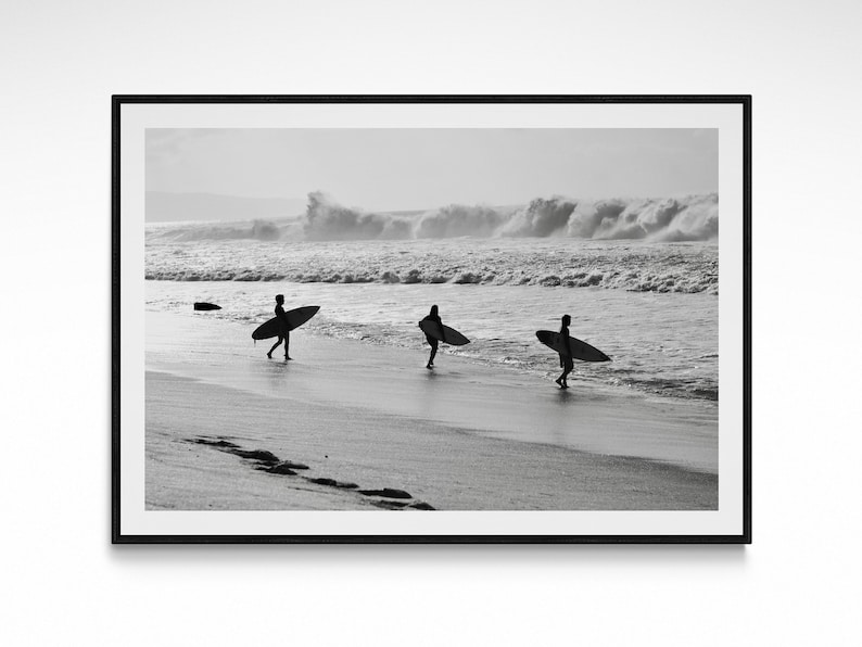 May include: Black and white photograph of three people walking on a beach with surfboards in hand. The people are silhouetted against the ocean and the waves crashing in the background.