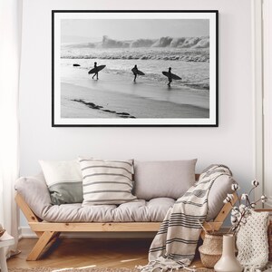 May include: Black and white photograph of three surfers walking on a beach with their surfboards. The photo is framed in a black frame and hanging on a white wall above a light gray couch.