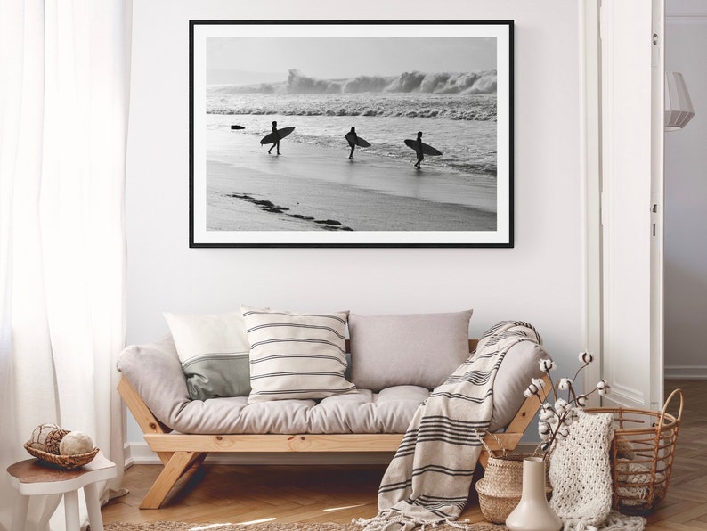 May include: Black and white photograph of three surfers walking on a beach with their surfboards. The photo is framed in a black frame and hanging on a white wall above a light gray couch.