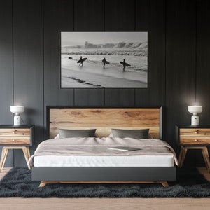 May include: A bedroom with a wooden bed frame, a black headboard, and a gray duvet. Two nightstands with lamps are on either side of the bed. A black and white photograph of surfers on a beach hangs above the headboard.