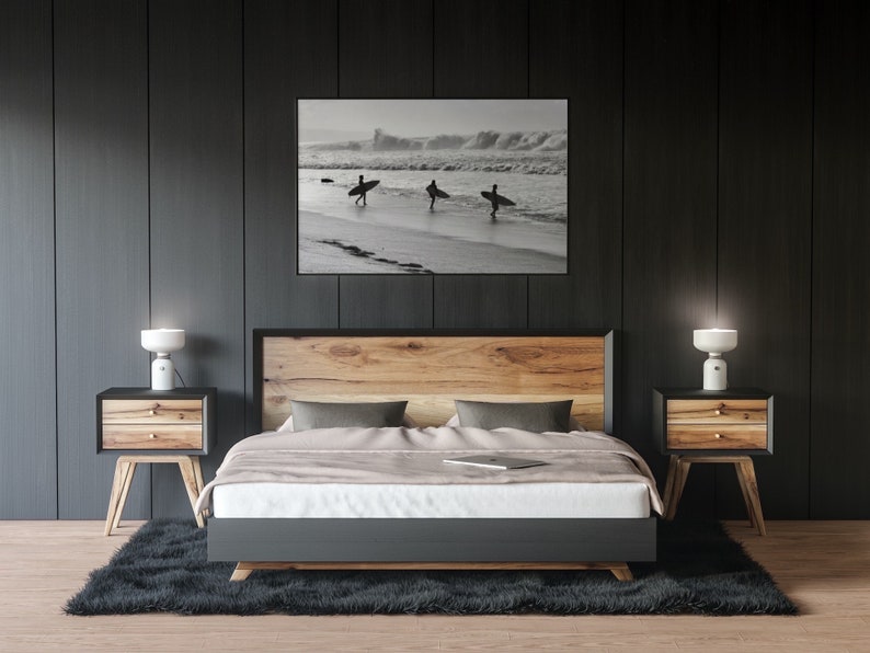 May include: A bedroom with a wooden bed frame, a black headboard, and a gray duvet. Two nightstands with lamps are on either side of the bed. A black and white photograph of surfers on a beach hangs above the headboard.