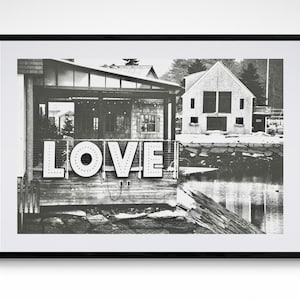 May include: Black and white photograph of a building with the word "LOVE" in large, white letters on the front. The building is located on a waterfront with a wooden dock and a body of water in the background.