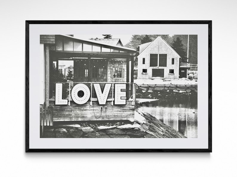 May include: Black and white photograph of a building with the word "LOVE" in large, white letters on the front. The building is located on a waterfront with a wooden dock and a body of water in the background.