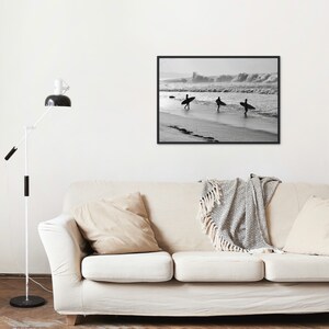 May include: Black and white photograph of three surfers walking on a beach with their surfboards. The surfers are silhouetted against the waves in the background.