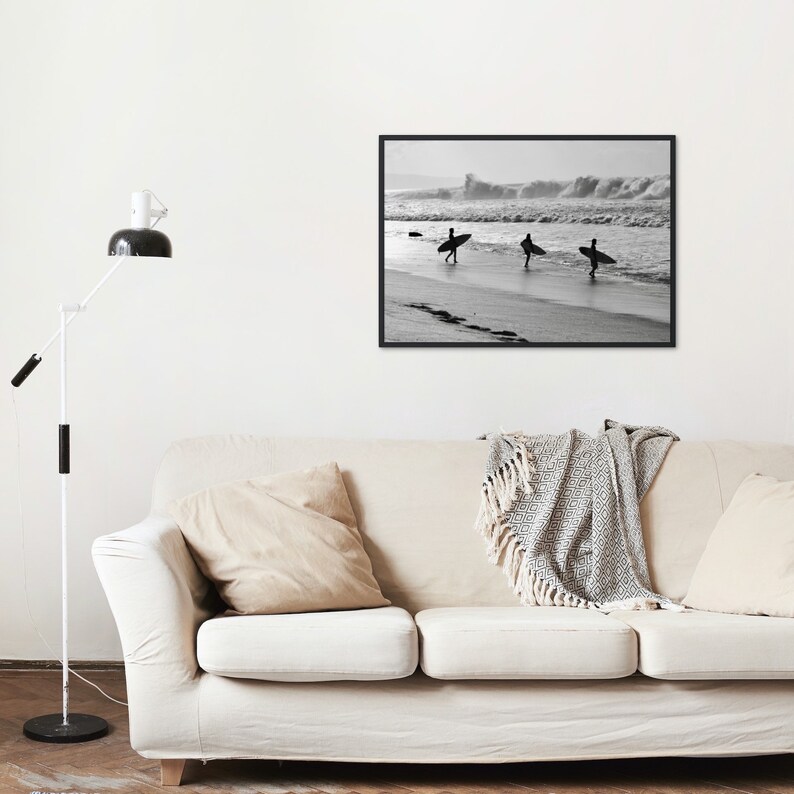May include: Black and white photograph of three surfers walking on a beach with their surfboards. The surfers are silhouetted against the waves in the background.