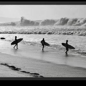 May include: Three people walk along a sandy beach carrying surfboards. They are silhouetted against a bright, cloudy sky and a choppy ocean.