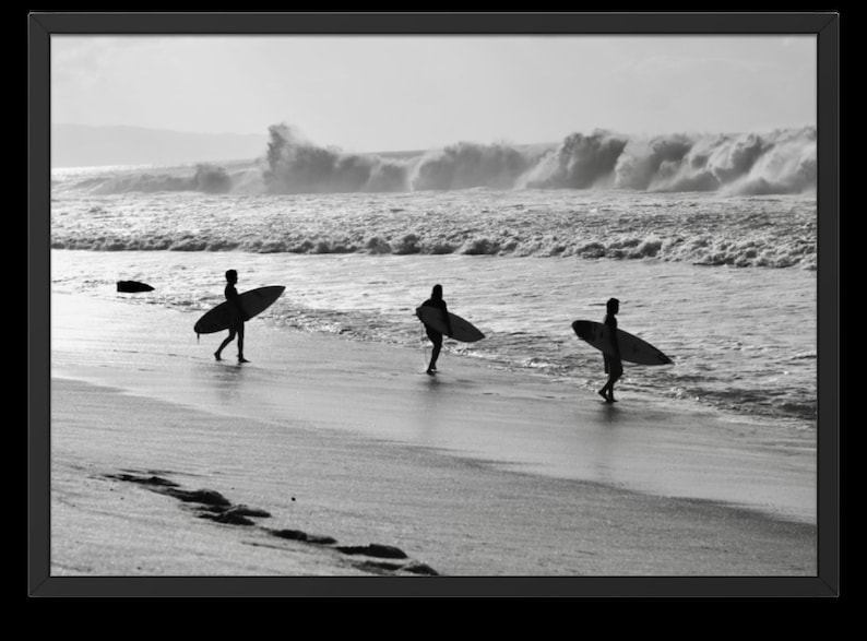 May include: Three people walk along a sandy beach carrying surfboards. They are silhouetted against a bright, cloudy sky and a choppy ocean.