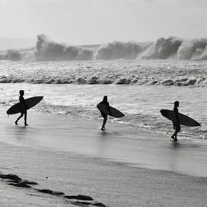 May include: Three people walk along a sandy beach carrying surfboards. The beach is empty except for the three people and the surfboards. The ocean is in the background with waves crashing in the distance.
