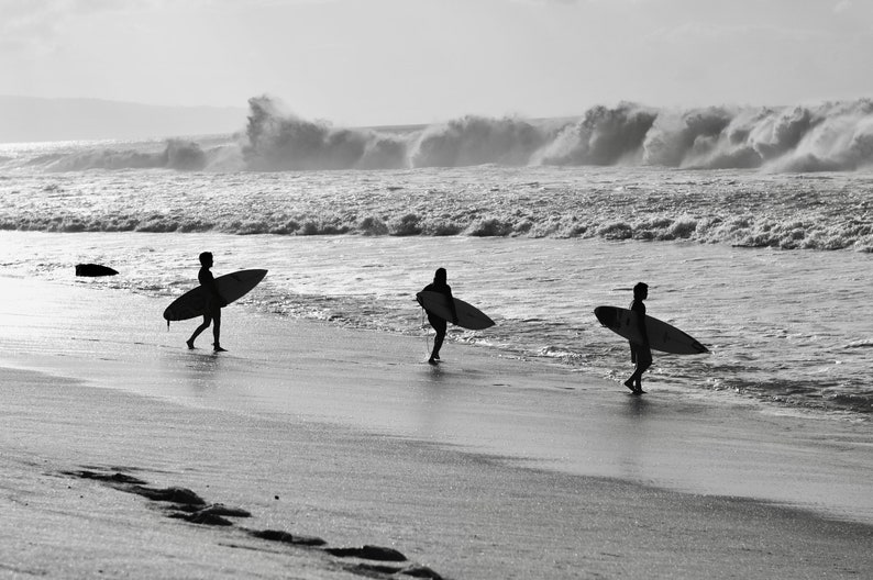May include: Three people walk along a sandy beach carrying surfboards. The beach is empty except for the three people and the surfboards. The ocean is in the background with waves crashing in the distance.