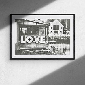 May include: Black and white photograph of a building with a sign that says "LOVE" in large, bold letters. The building is located on a waterfront with a small body of water in the foreground.