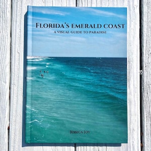 May include: A book titled "Florida's Emerald Coast" with a visual guide to paradise. The cover features a vibrant image of the ocean with turquoise water and a blue sky. The author's name, Jessica Joy, is at the bottom.