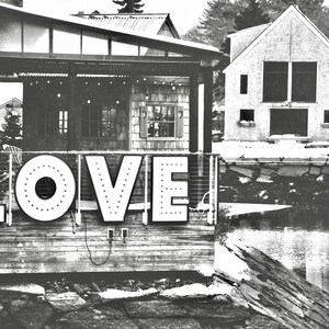 May include: A black and white photo of a wooden building with a sign that says "LOVE" in large, white letters. The sign is lit up with small white lights. The building is located on a waterfront with a rocky shore and a body of water in the background.