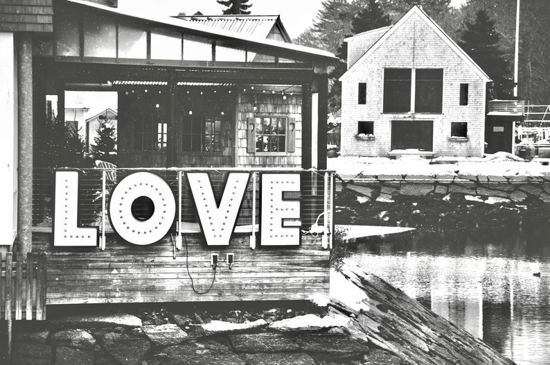 May include: A black and white photo of a wooden building with a sign that says "LOVE" in large, white letters. The sign is lit up with small white lights. The building is located on a waterfront with a rocky shore and a body of water in the background.