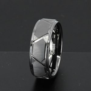 May include: A silver tungsten carbide ring with a geometric design. The ring has a brushed finish and the words "Tungsten Carbide" are engraved on the side.